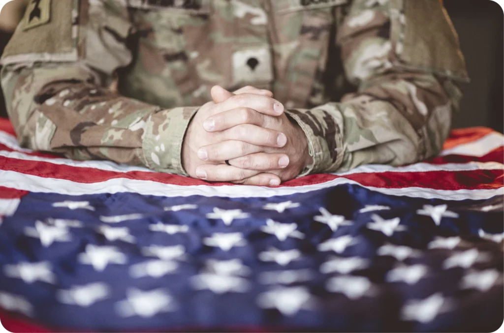 Service member in camouflage uniform with hands clasped over an American flag
