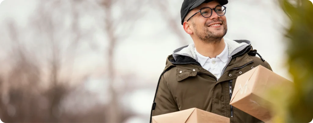 Smiling delivery driver in a cap and glasses carrying plain cardboard boxes