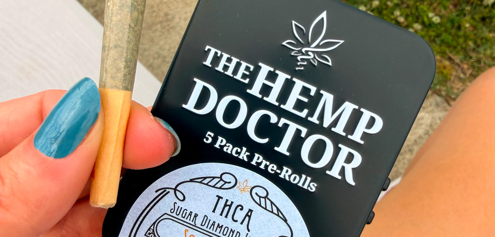 A hand holding a pre-roll in front of The Hemp Doctor 5-pack packaging
